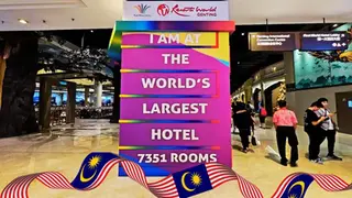 Colorful sign in a hotel lobby reads "I am at the world's largest hotel, 7351 rooms." People walk by under signs and decor. Malaysian flag displayed.