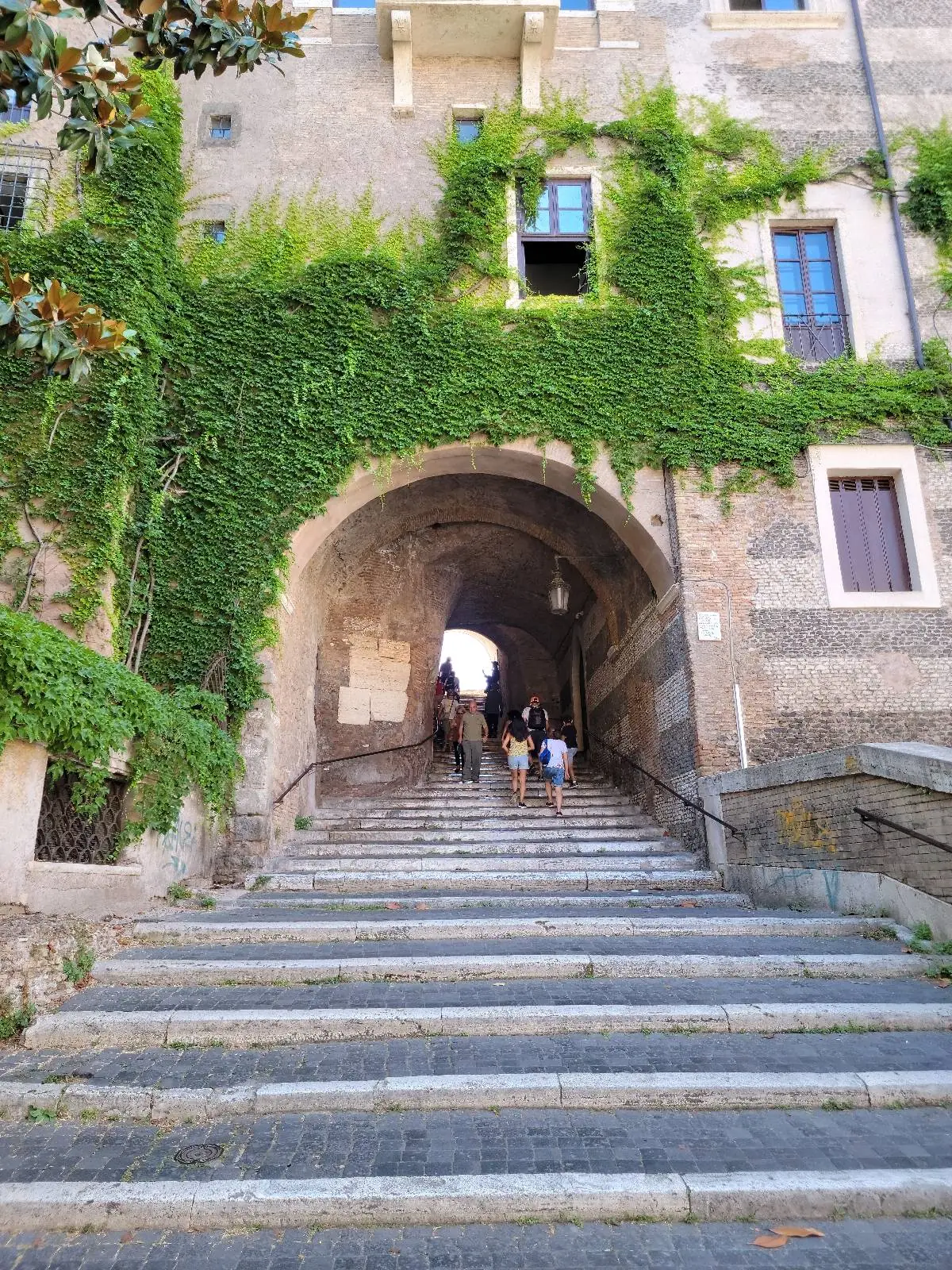 Stone steps lead up to an arched passageway in an old building, partly covered with green ivy. Sunlight brightens the entrance and a few people are visible walking under the archway.