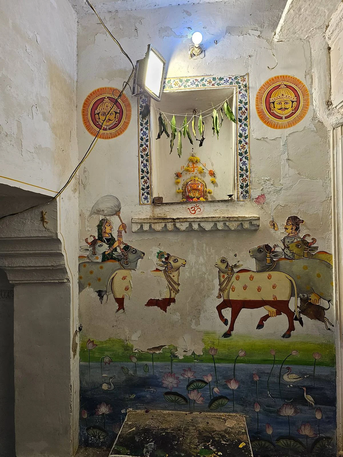 A colorful mural on a wall depicts cows, figures, and decorative patterns. Above the mural, there is a small shrine with a statue and religious symbols, all lit by a single bulb. The walls are weathered and slightly cracked.
