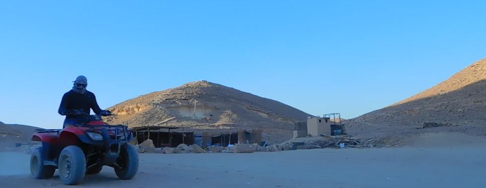 A person rides an ATV on a sandy landscape under a clear blue sky. Two hills are visible in the background, along with a small building structure. The scene conveys a sense of adventure in a desert setting.