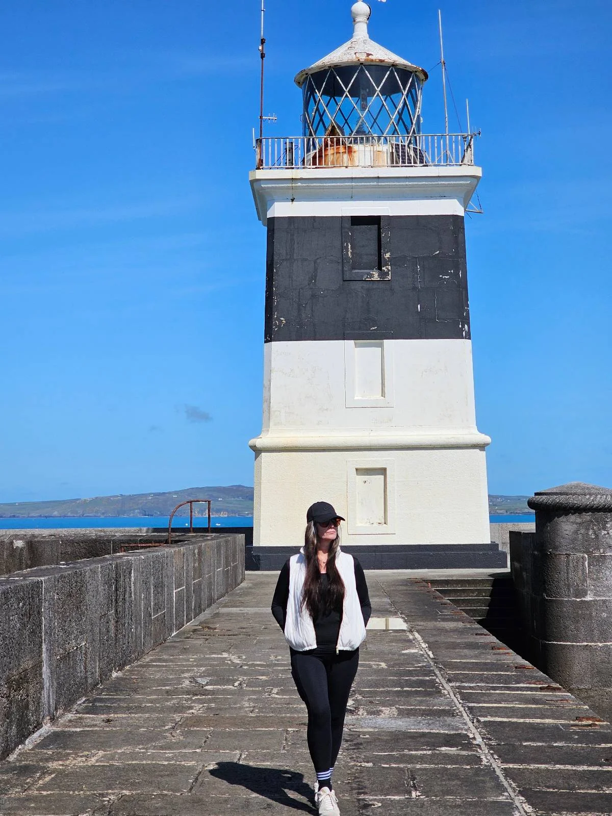 A woman walking towards the camera on a pier with a small white and black lighthouse in the background under a clear blue sky.