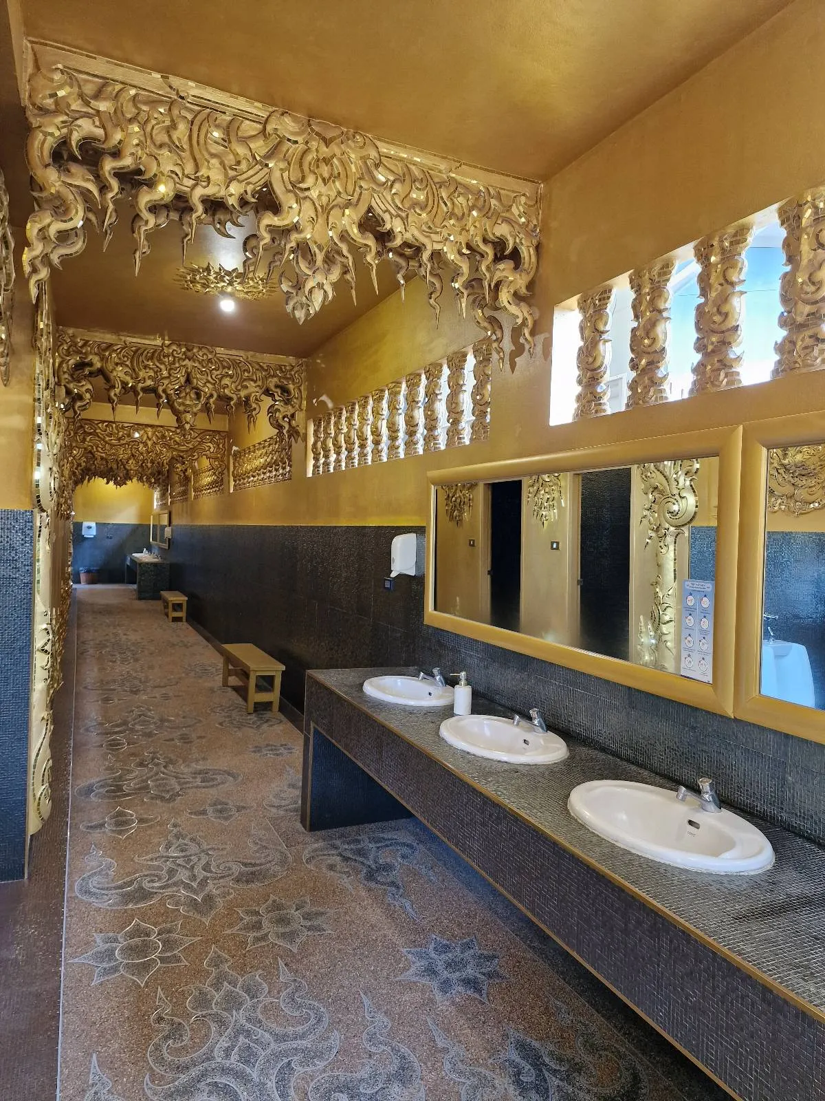 A restroom with ornate golden decorations on the ceiling and walls. There are three white sinks set in a dark counter, above which is a large mirror. The floor has an intricate pattern, and light enters through windows on the right.
