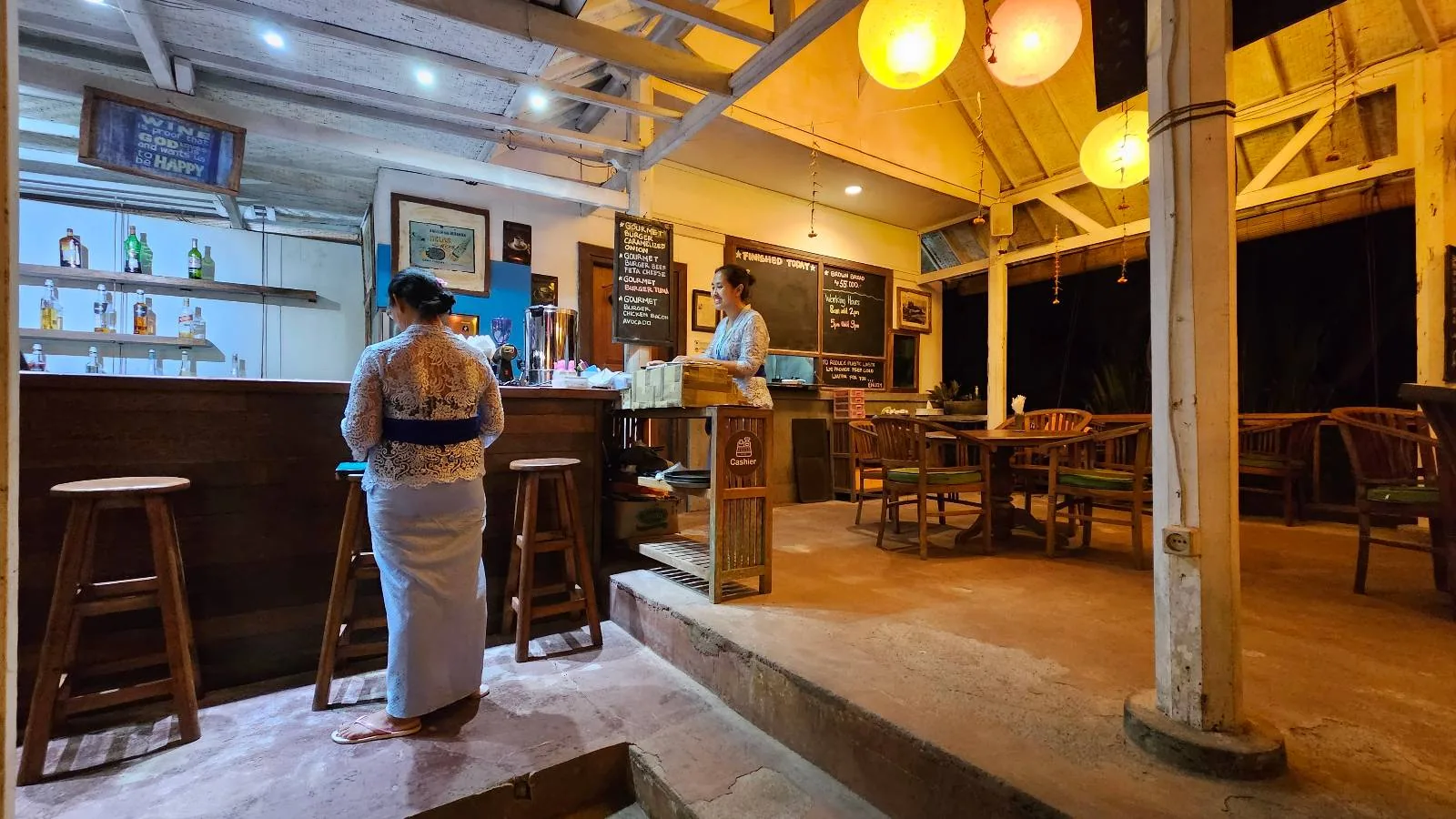 A warmly lit restaurant with a wooden bar and stools. A person in traditional attire stands near the bar, and another person is behind it. Round lanterns hang from the ceiling, and tables with chairs are set up in the background.