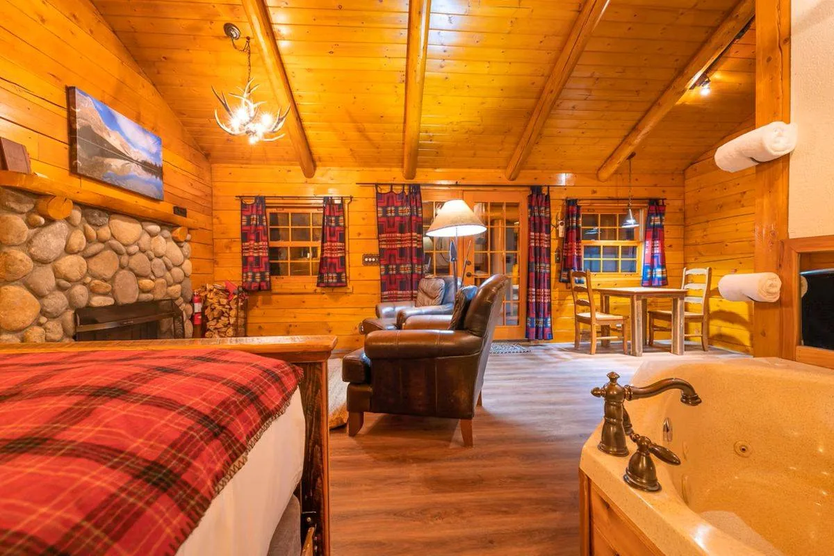 Cozy cabin interior with wooden walls and ceiling. Features a stone fireplace, plaid-covered bed, and leather armchair. A chandelier hangs above, and dining table with chairs is near the window. A bathtub with bronze fixtures is in the foreground.
