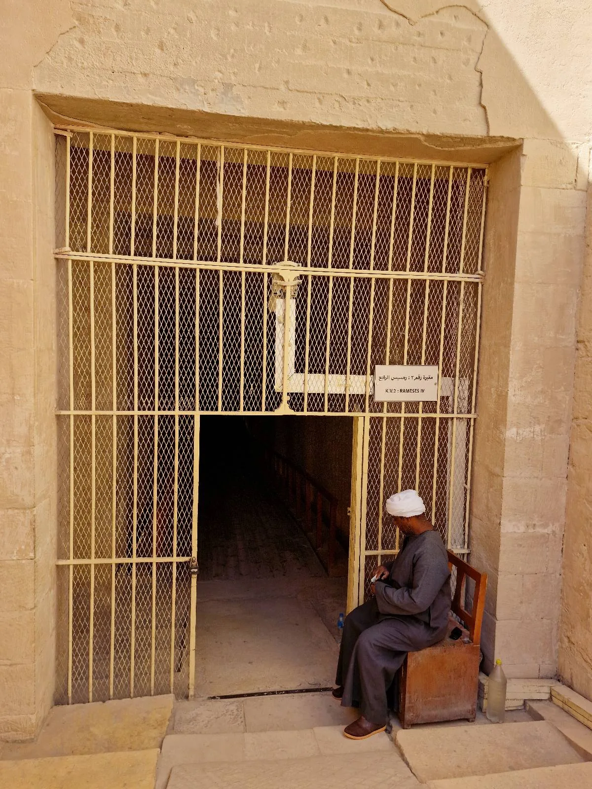 A person sits on a bench outside a gated entrance to a building. The sign on the gate reads "KV2 Ramses IV".