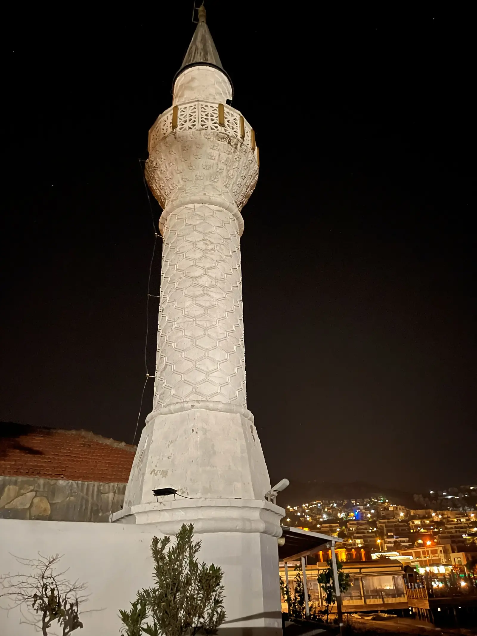 A tall, white minaret stands against a night sky, with city lights visible in the background.