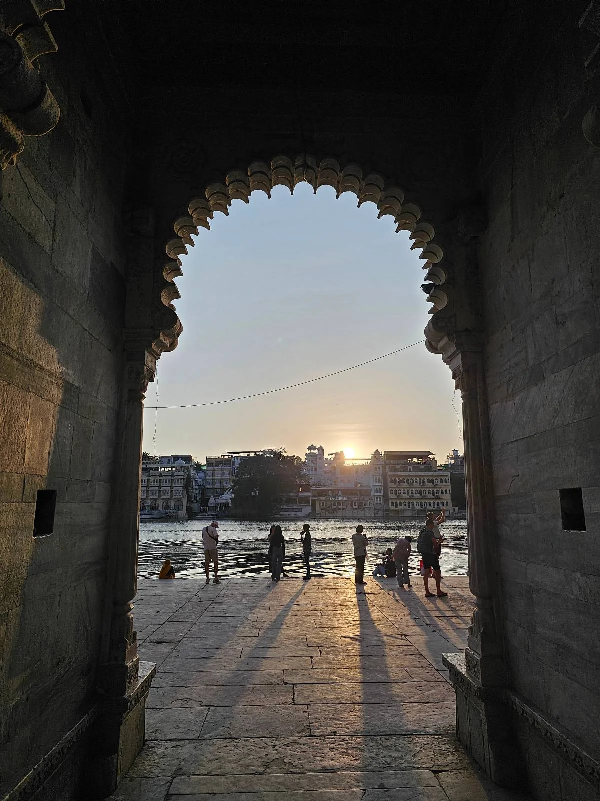 A sunset view through a large arched stone doorway, with several people silhouetted against the light, overlooking a body of water and buildings in the distance.