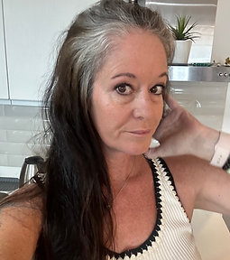 A woman with long dark hair and streaks of gray poses indoors, touching her hair. She wears a sleeveless top with black trim, and a light-colored watch. A plant and kitchen items are visible in the background.