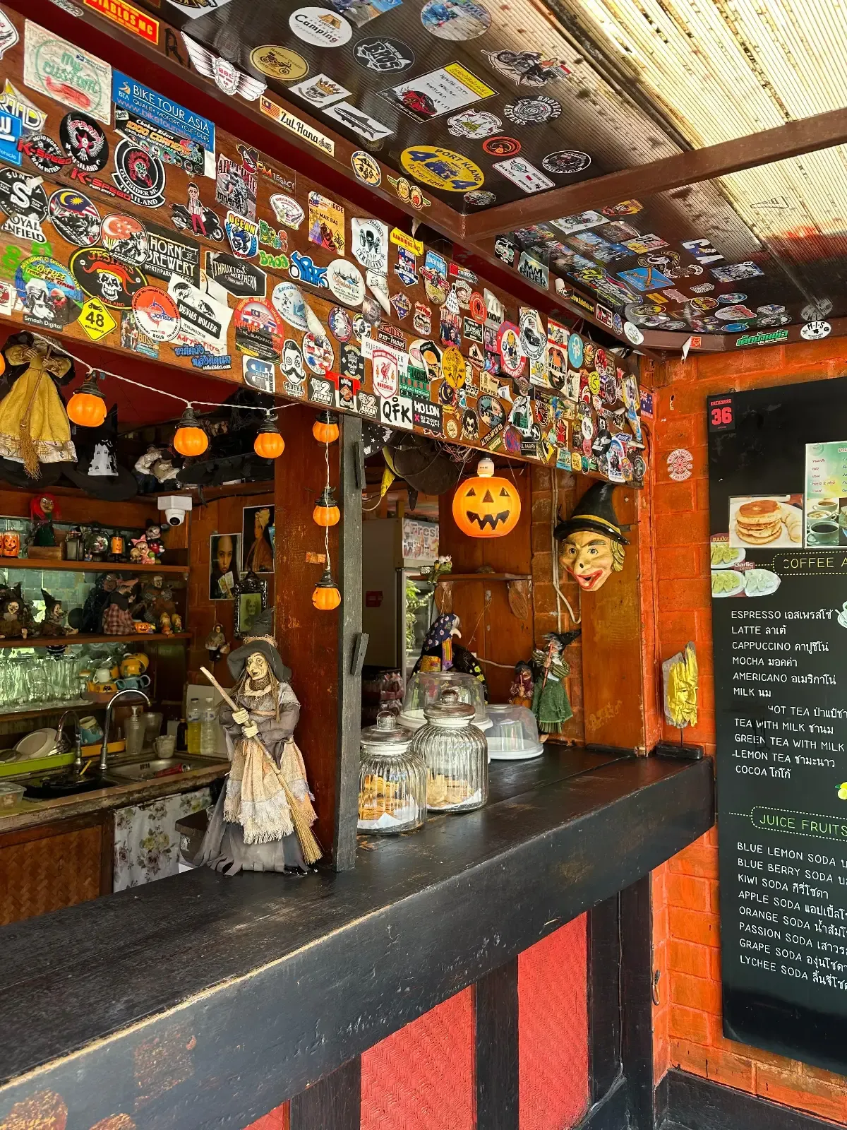 A small, colorful café counter decorated with stickers, Halloween ornaments, and jars. A pumpkin and scarecrow hang above the counter, and a chalkboard menu is visible on the right side.