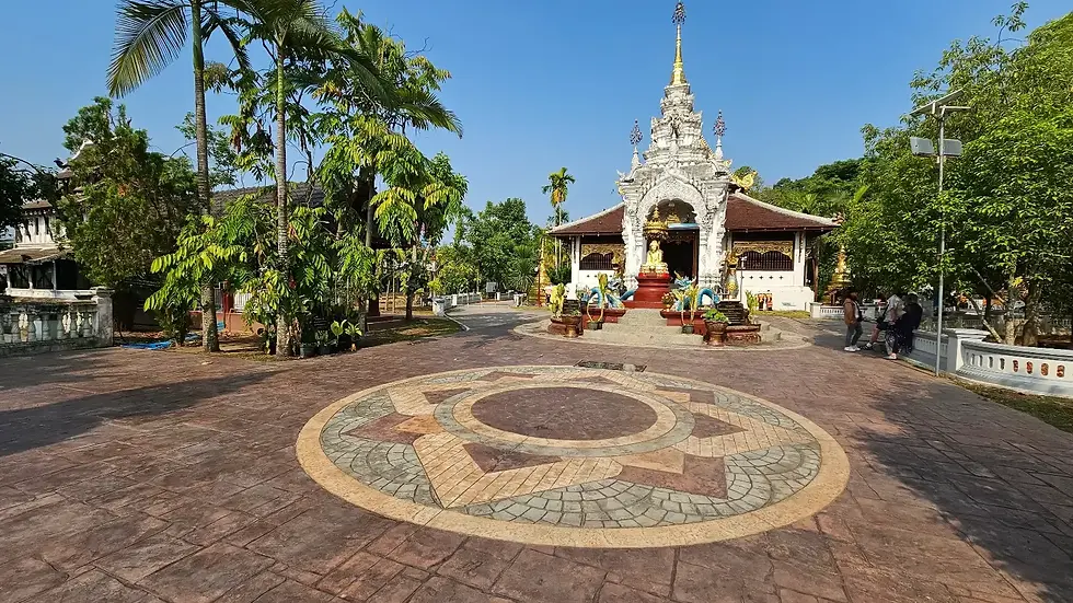 Ornate white temple with gold accents under clear blue sky, surrounded by lush green trees. Intricate circular pattern on plaza in foreground.