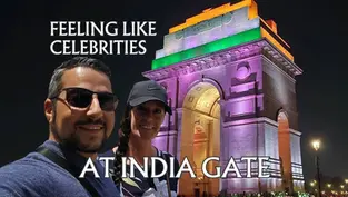 Two people smiling in front of a brightly lit India Gate at night. Text reads "Feeling like celebrities at India Gate."
