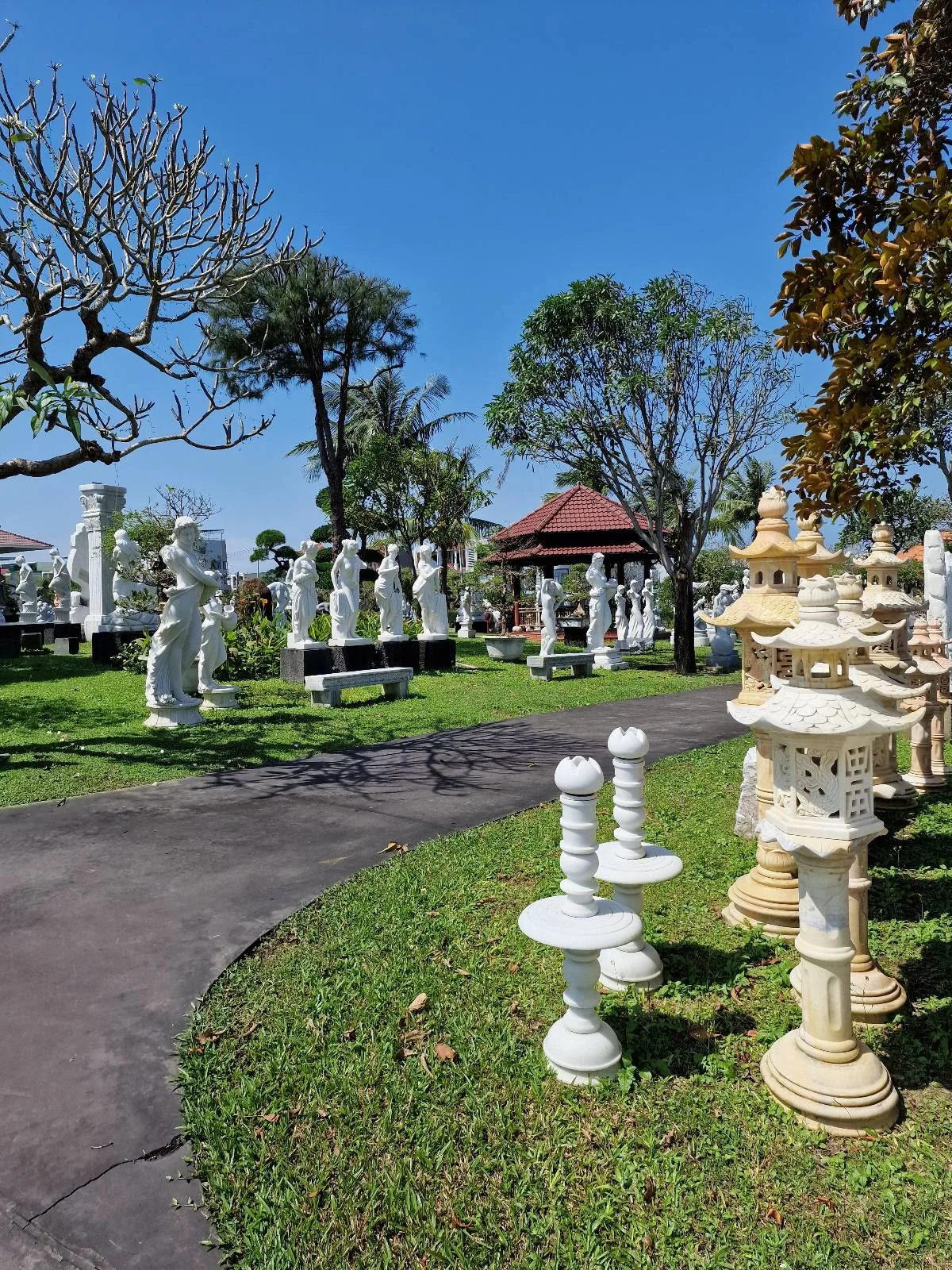 A garden scene with large, white statues, bases and sculptures on grass. Trees and a gazebo are in the background under a clear blue sky.