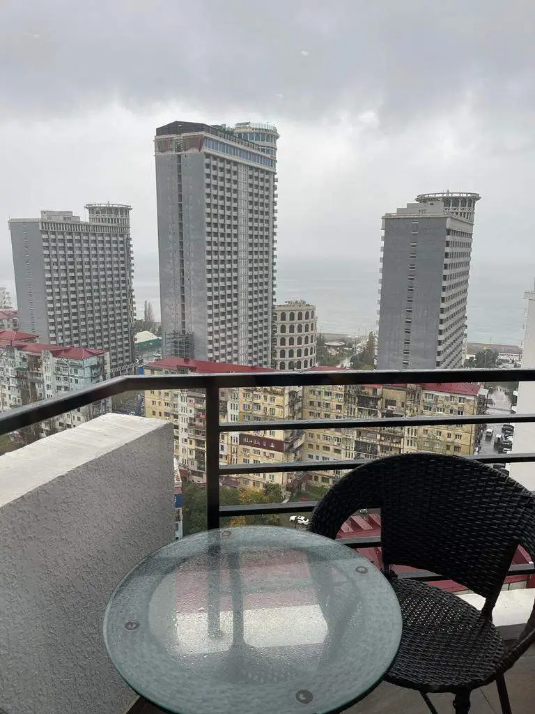 A small balcony with a glass table and a chair overlooking a several skyscrapers in a city.