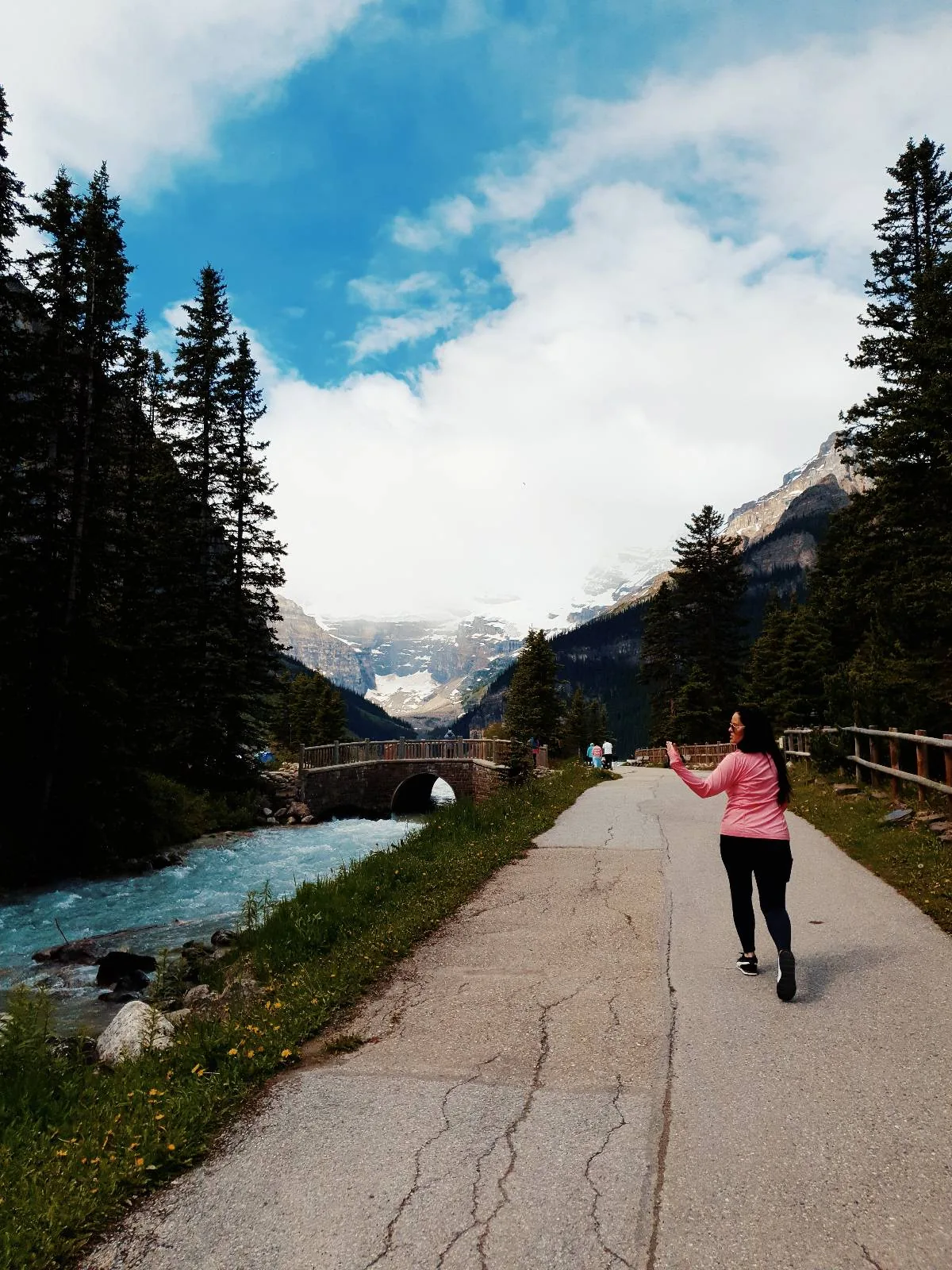 A woman in a pink jacket walks on a paved path beside a river, surrounded by pine trees and mountains under a partly cloudy sky.