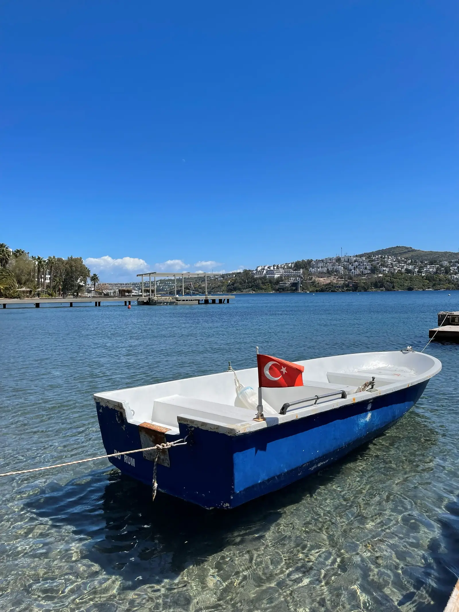 A blue and white rowboat with a Turkish flag floats on clear water, with a distant view of hills and a bright sky.