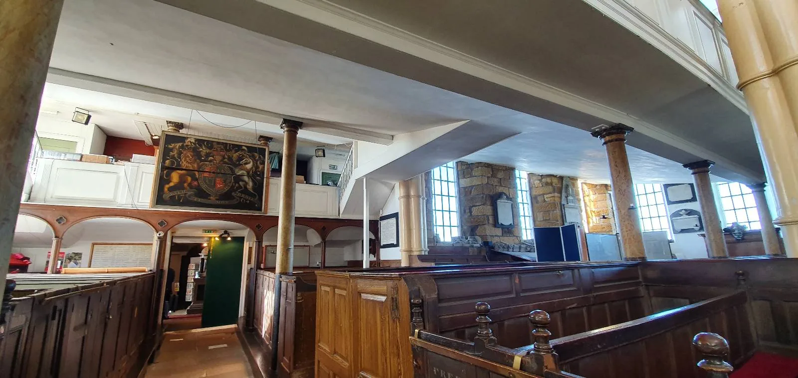 A view of the interior of a historic church, featuring wooden pews, columns, large windows, and religious paintings on the walls under a white ceiling.