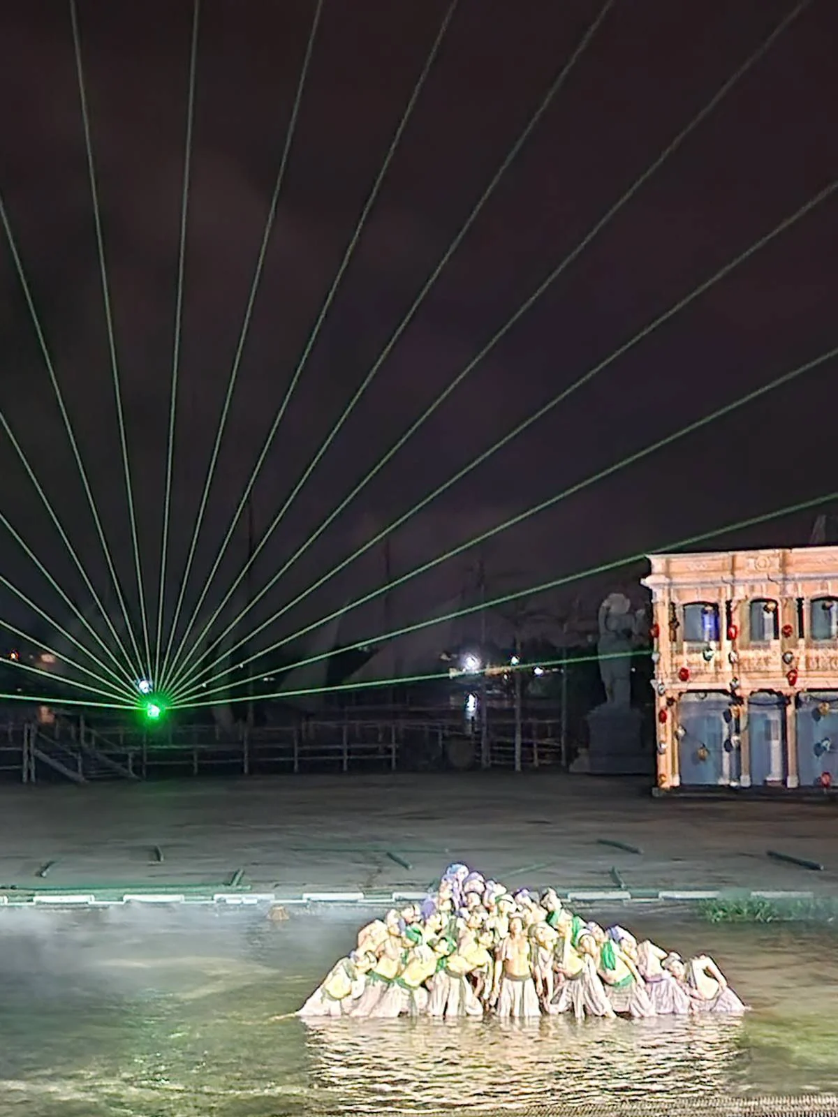 Nighttime theatrical performance with actors dressed in period costumes on a river stage, bathed in green laser lights and a foggy atmosphere, with an old-style building in the background.