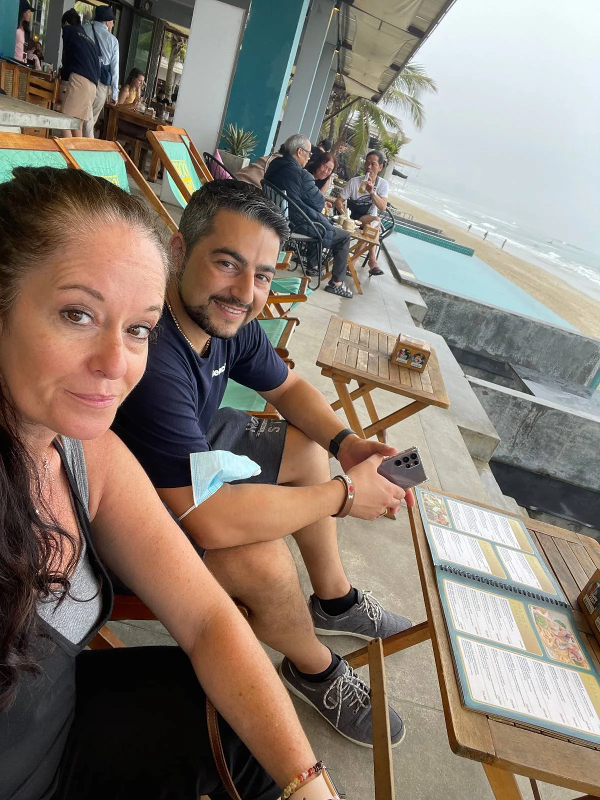 A woman and a man sit on a patio at a seaside cafe. They are holding menus and smiling at the camera. The patio overlooks a beach with waves and cloudy skies in the background. Others are seated at tables nearby.