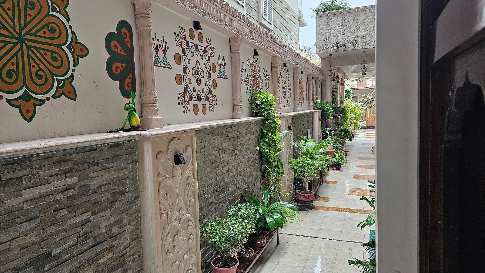 A narrow outdoor corridor with potted plants lining one side, decorative wall art, stone textures, and sunlight coming in from an open gate at the far end.