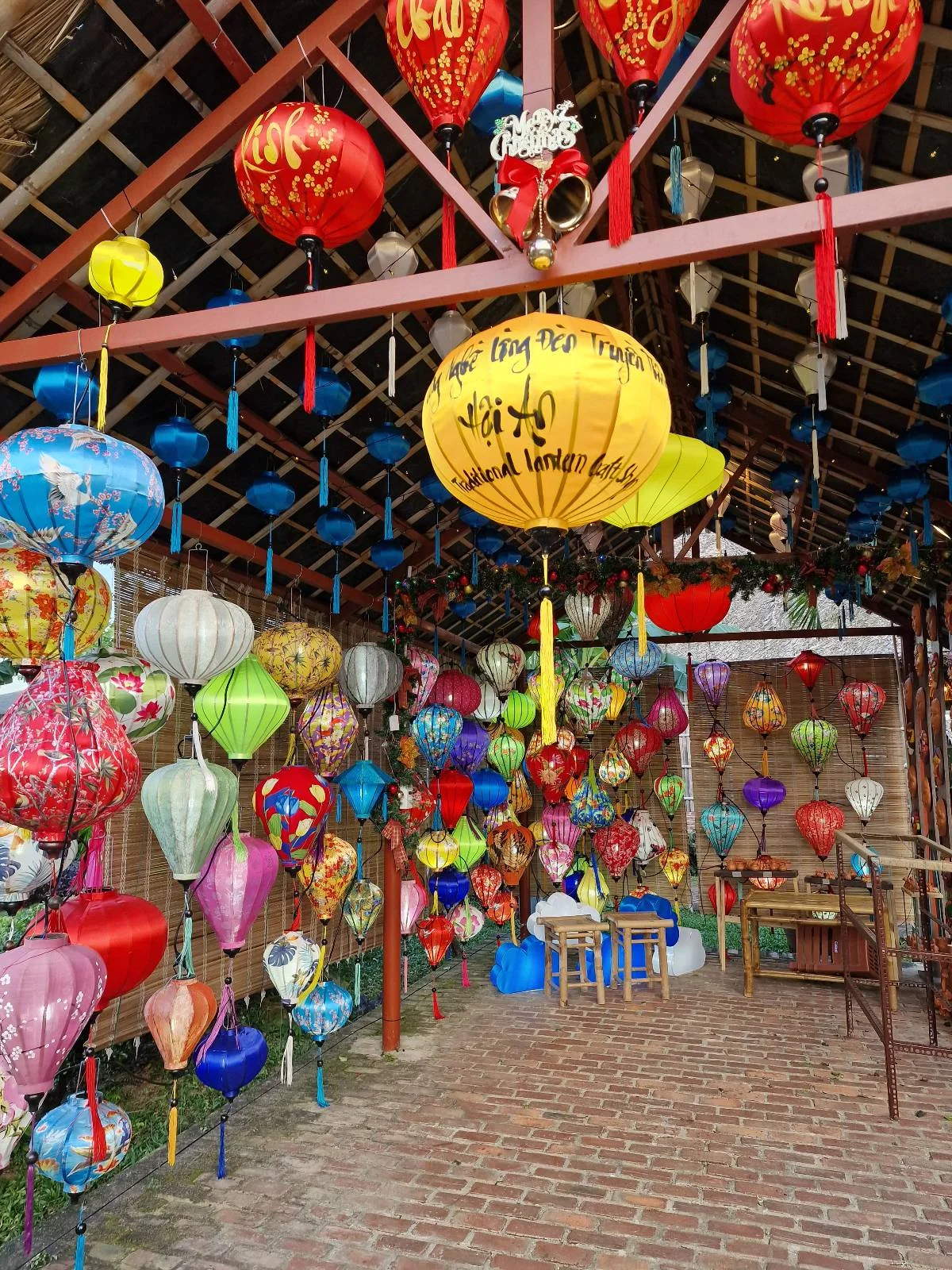 A colorful display of variously shaped and sized lanterns hanging from the ceiling of a covered outdoor area with brick flooring and walls, creating a vibrant and festive atmosphere.