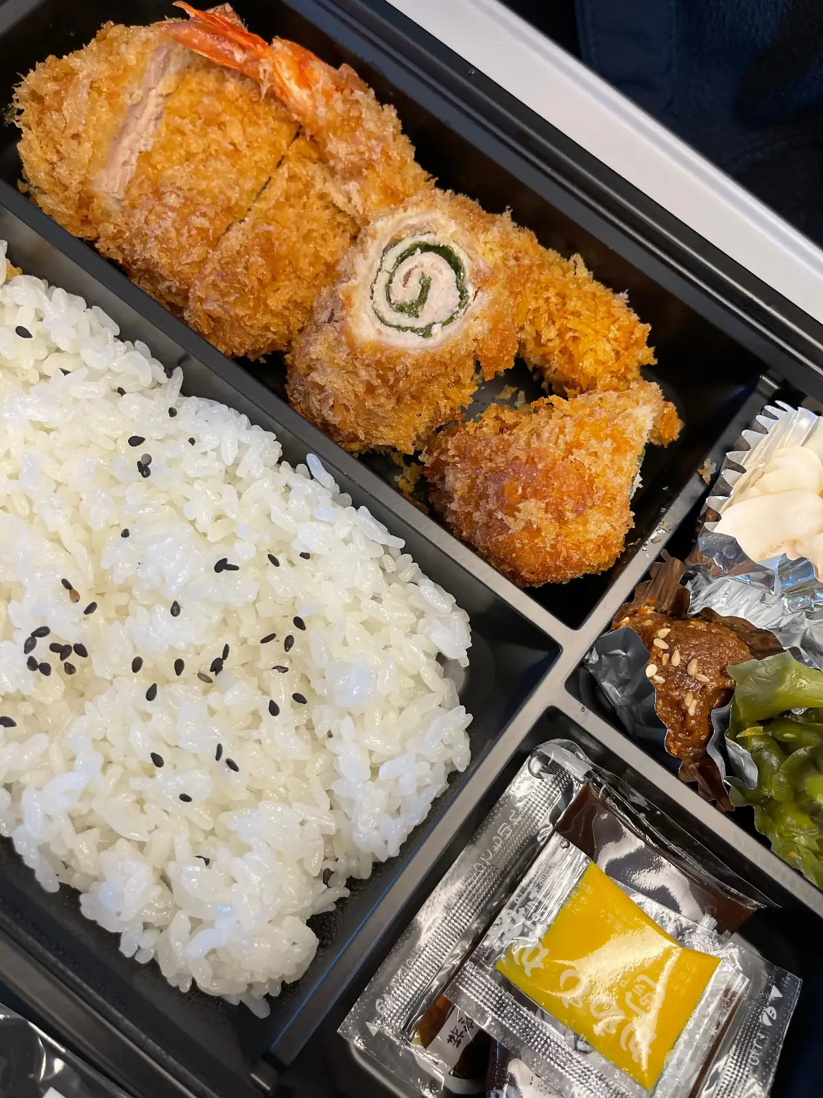 A bento box with white rice topped with black sesame seeds, breaded fried chicken pieces, a rolled cutlet with filling, a compartment of vegetables, and a packet of yellow sauce.