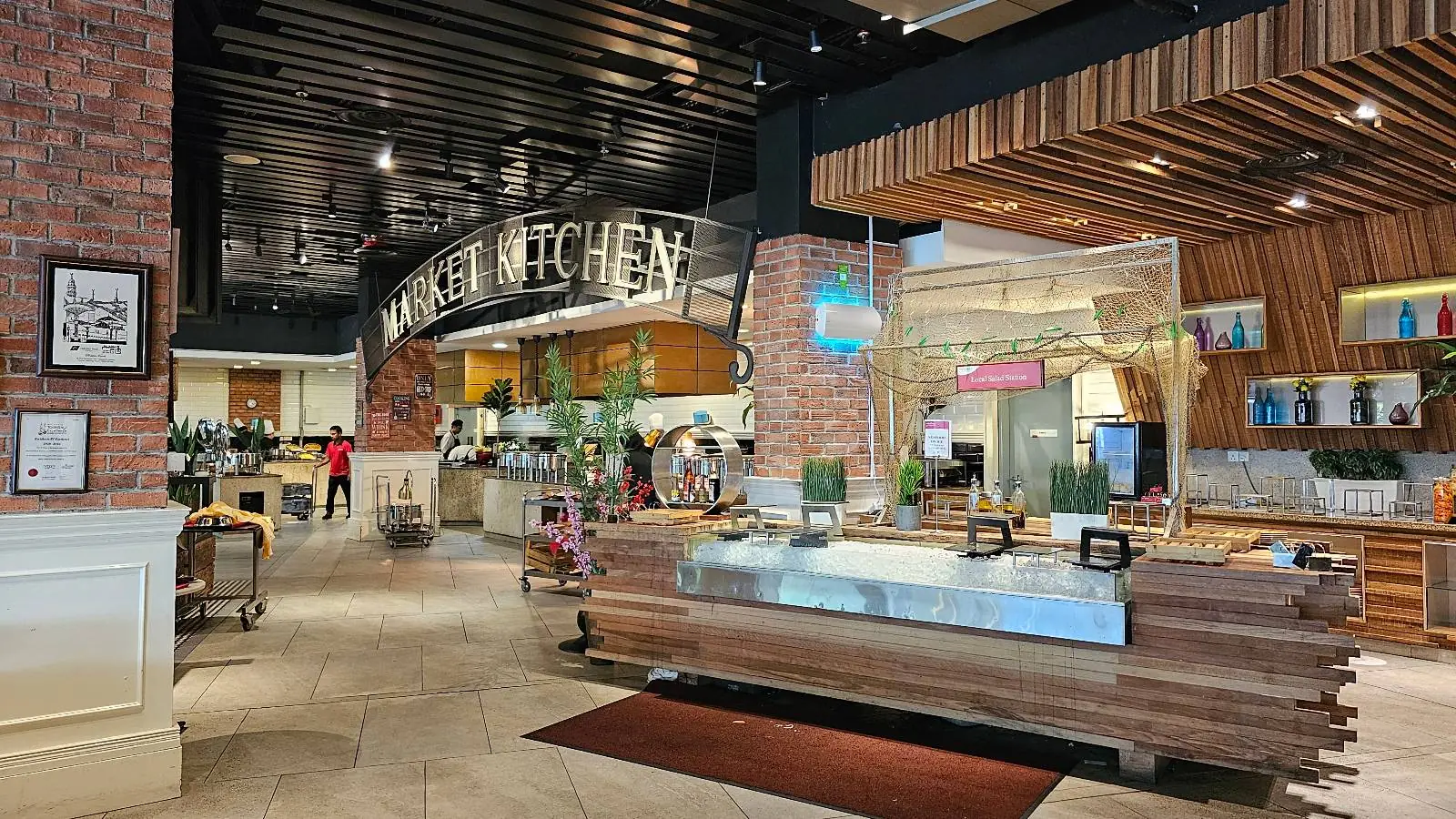 A bustling food court features a variety of food stalls with a sign reading "KITCHEN" above. The area has a modern design with exposed brick walls and wooden accents. Shoppers with carts can be seen in the background, and the setting is brightly lit and inviting.