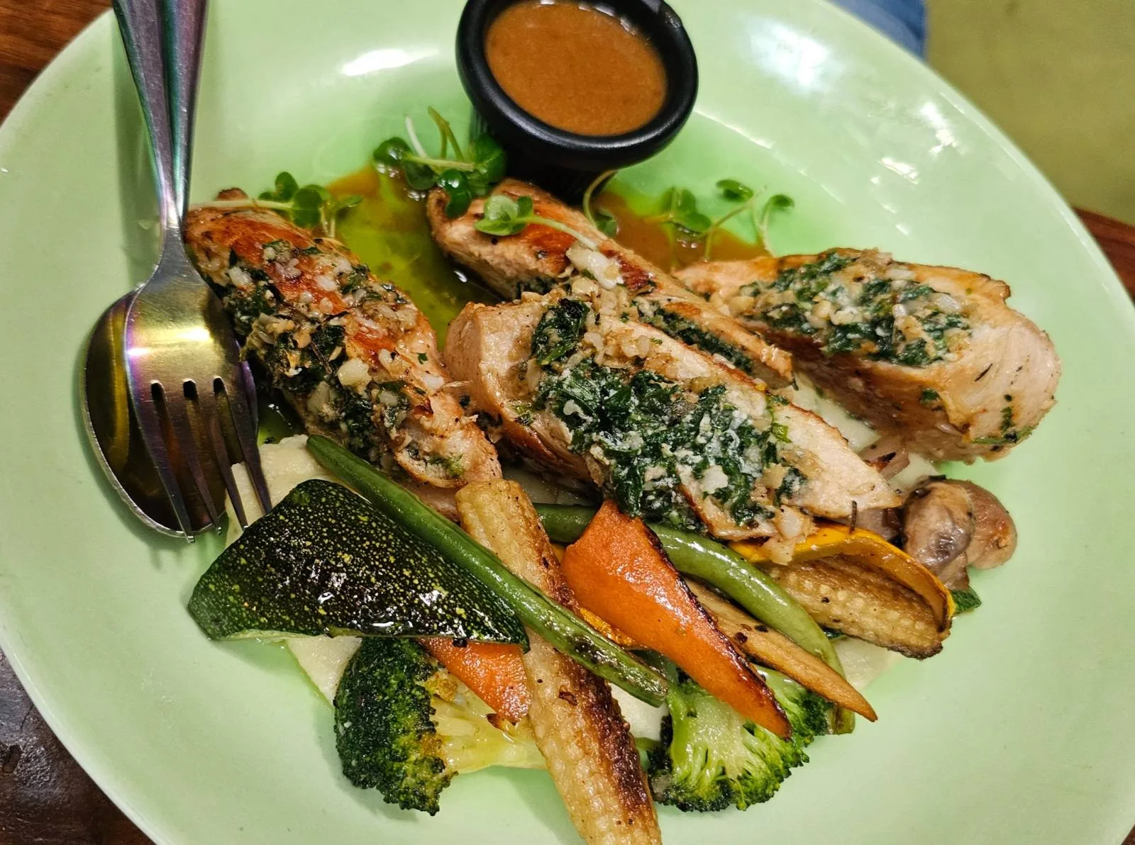 A plate of grilled chicken topped with herbs and spices, served with a side of sautéed vegetables including broccoli, zucchini, carrots, and baby corn. A small container of sauce is on the plate. Fork and spoon are placed on the side.
