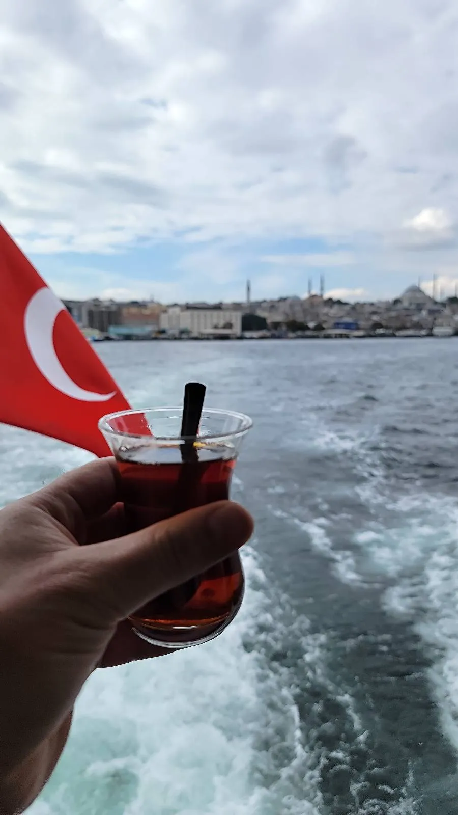 A person holds a glass of tea with a straw against the backdrop of a boat's wake, a Turkish flag, and a city skyline under a cloudy sky.