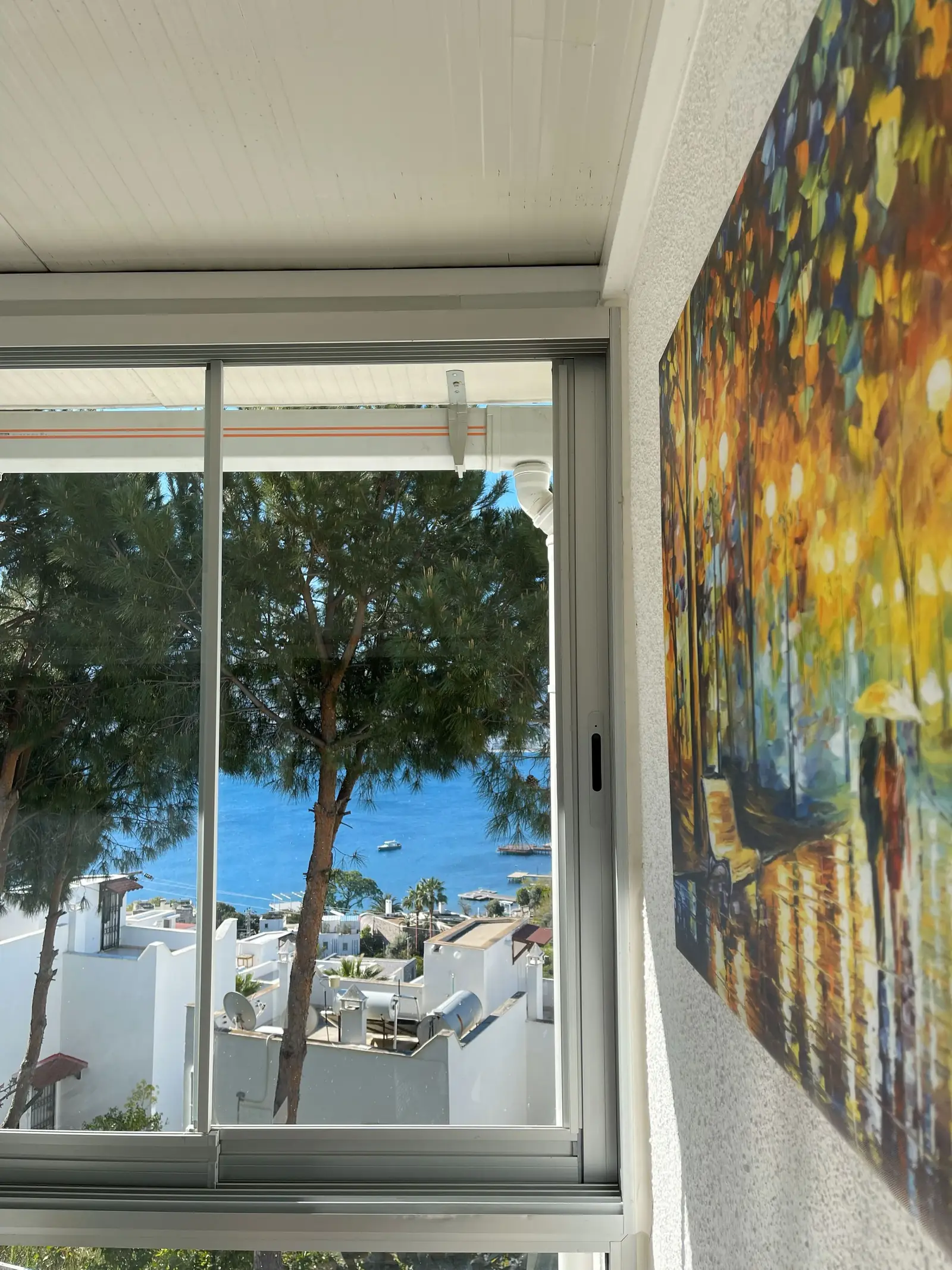 View from an indoor space with a sliding glass door showing a seaside scene with trees and boats. A colorful painting hangs on the wall.