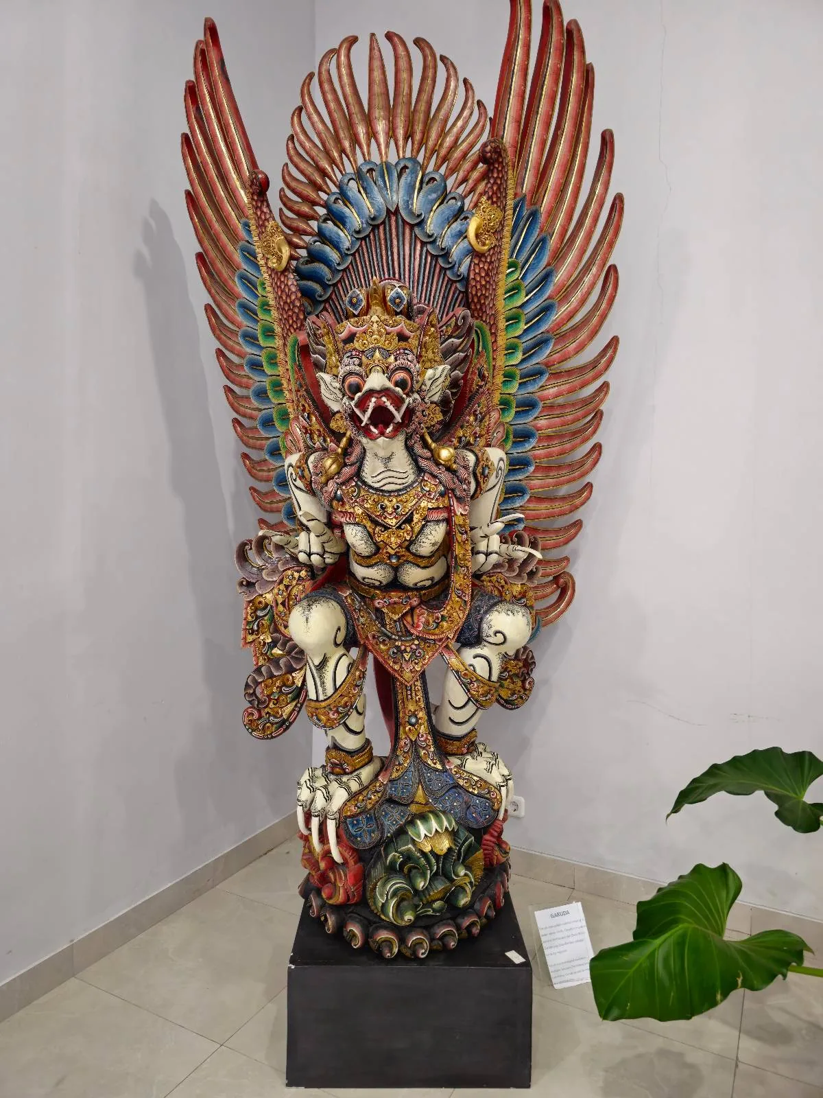 A colorful, intricately detailed statue of Garuda with outstretched wings stands on a black pedestal in a corner, next to a green plant. The Garuda has a fierce expression and ornate decorations.