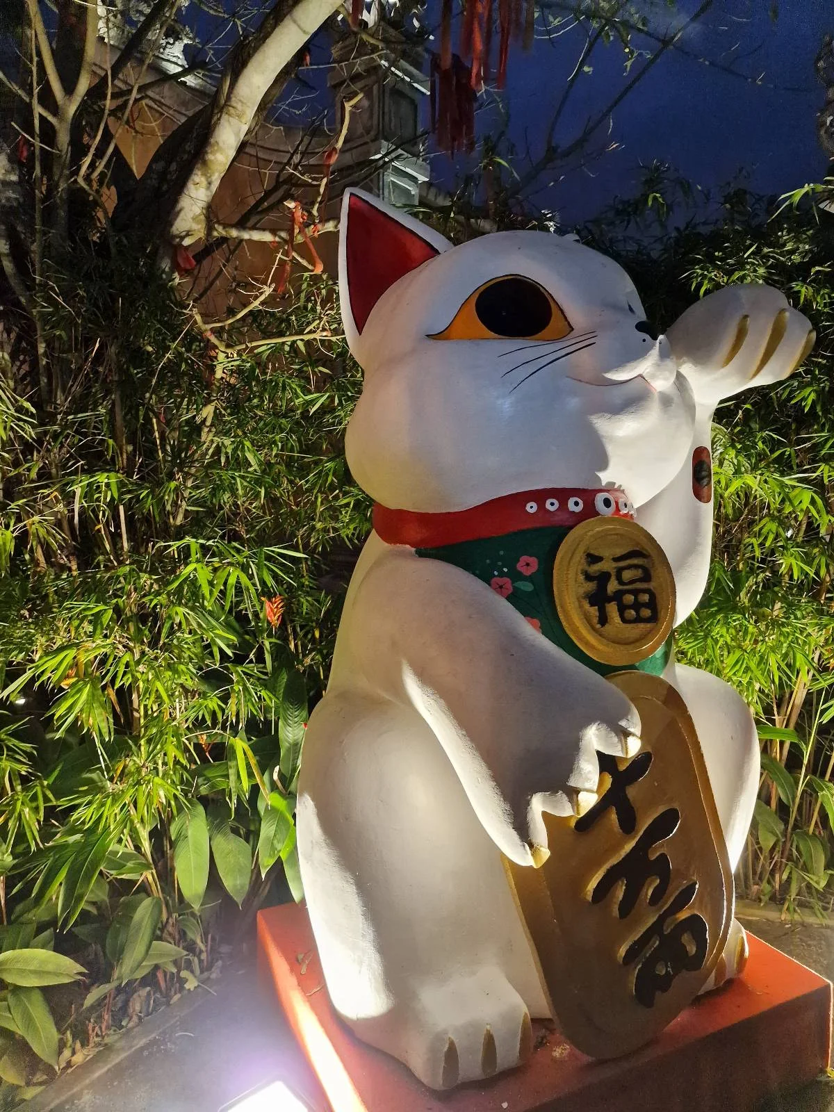 Large maneki-neko (lucky cat) statue illuminated at night, set among dense green foliage, holding a gold coin with Japanese characters.