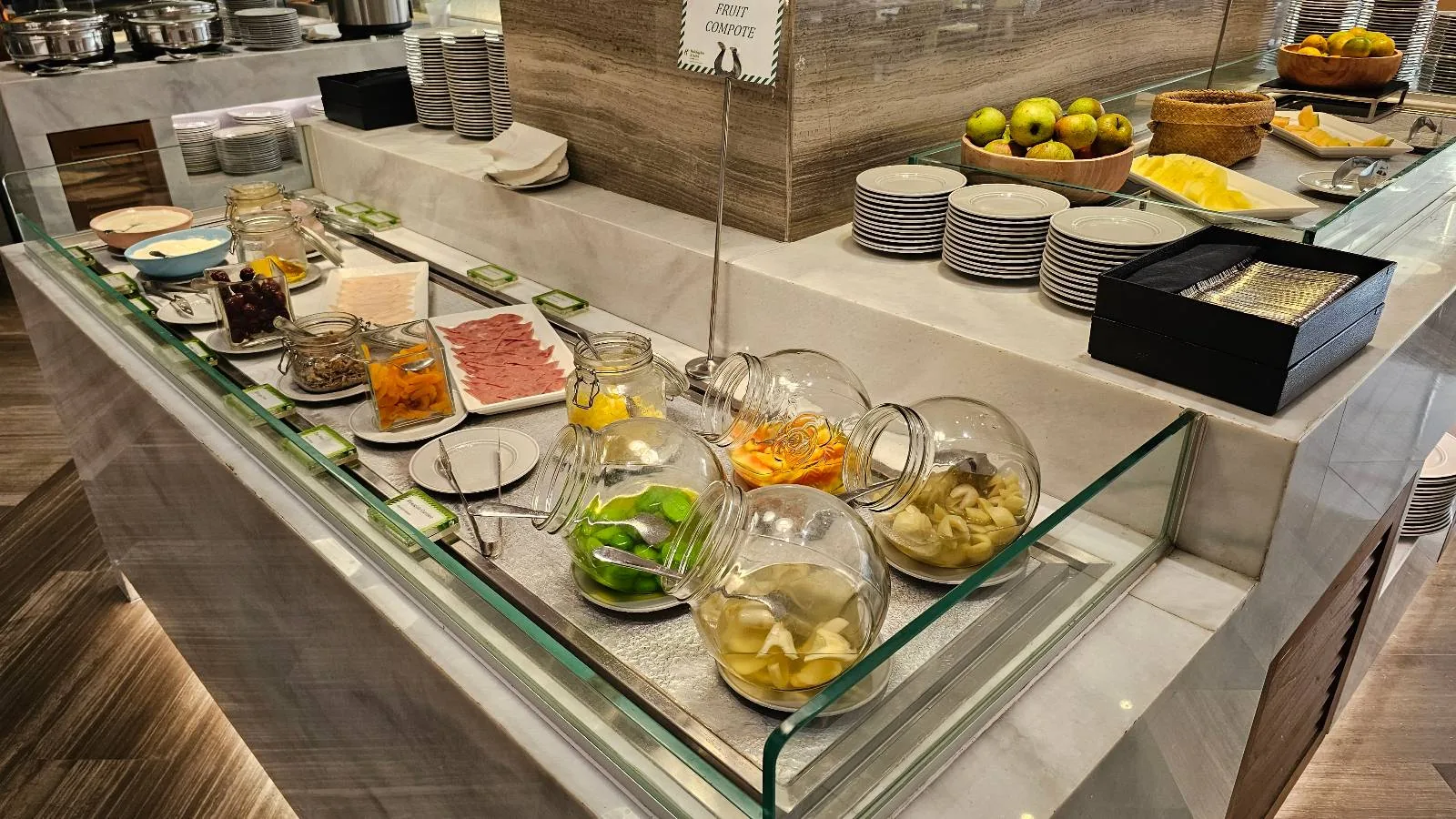 A hotel buffet counter displays various breakfast items, including sliced meats, cheeses, vegetables in glass jars, plates stacked in the background, and a bowl of fruit, all arranged neatly on a marble surface.