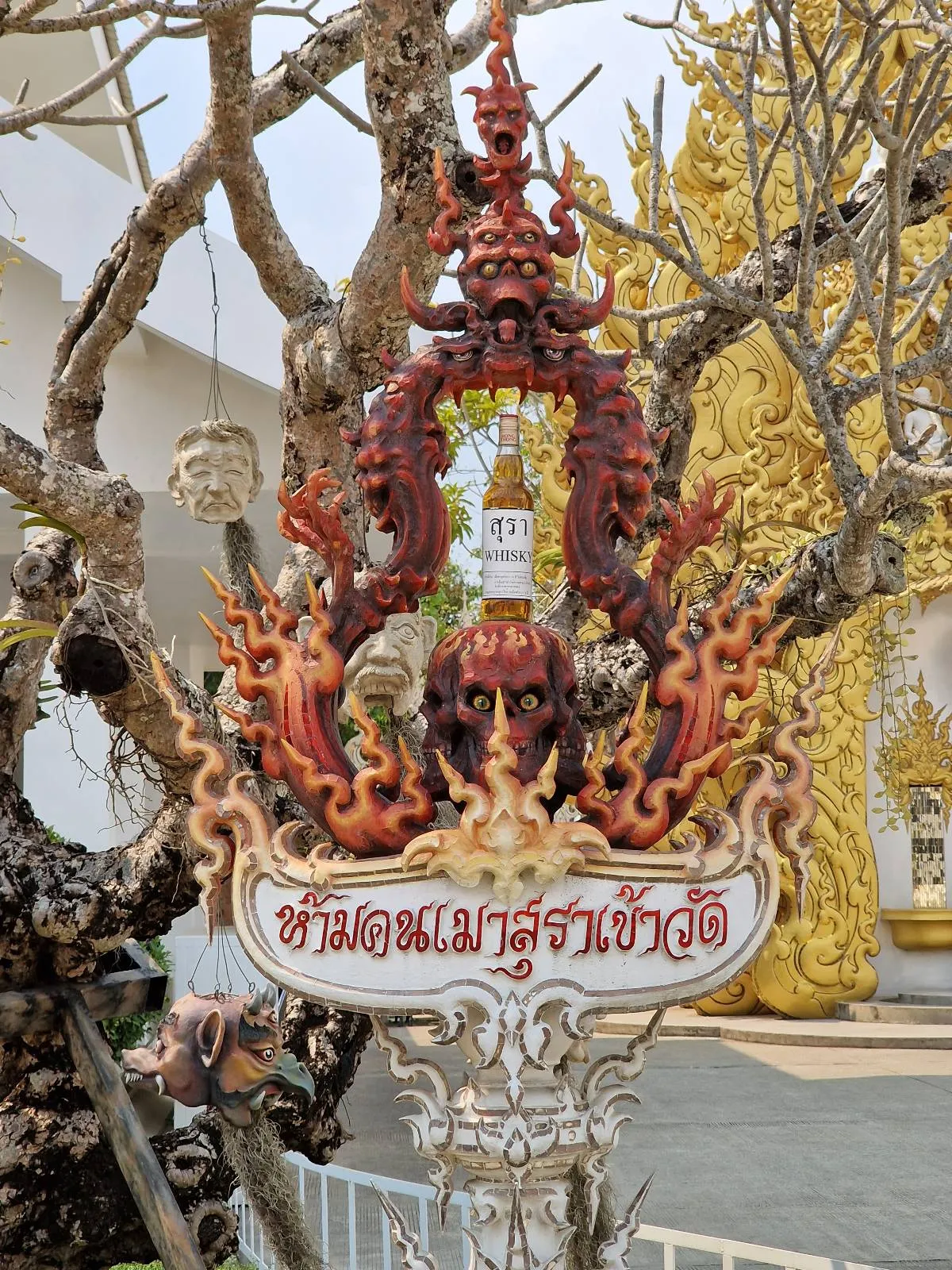 Ornate, fiery sculpture stands amidst tree branches in a temple setting, featuring intricate flaming details and Thai script on a base. Golden architectural elements are visible in the background.