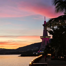 A tower with pink and purple lights stands by the water at sunset, with palm trees on the right and a lit path leading up to it.