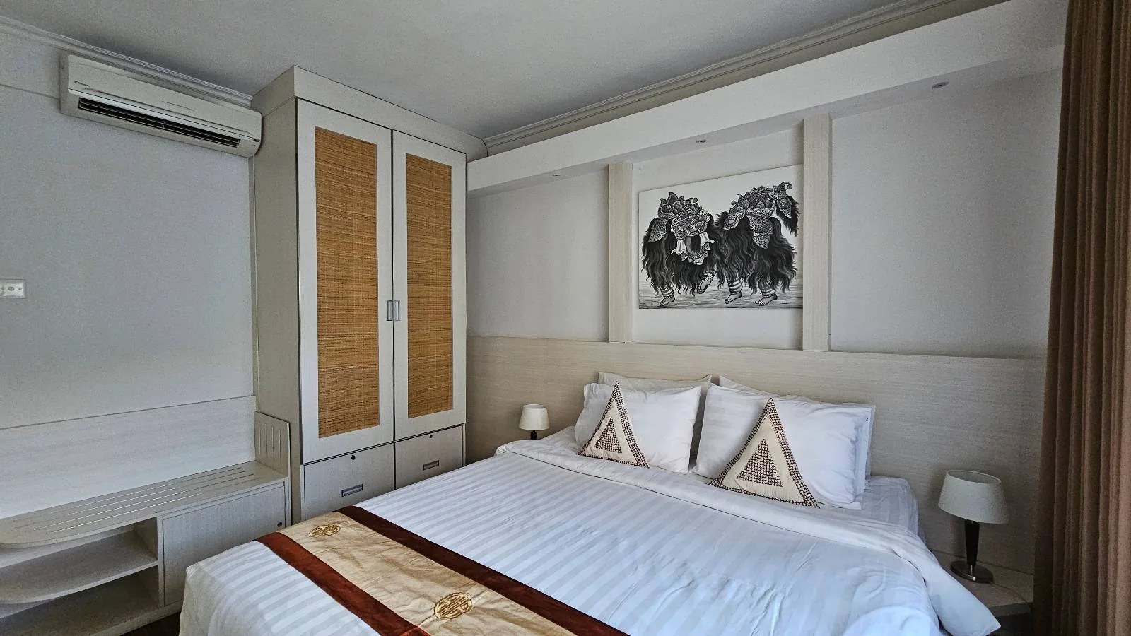 A neatly made bed with white linens and decorative pillows is in a modern bedroom. Above the bed is a black-and-white artwork of two lions. The room has built-in cupboards, two bedside lamps, and an air conditioner.