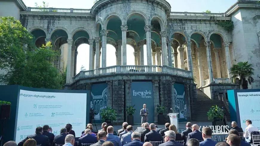 A person delivers a speech to an audience at an outdoor event with classical architecture in the background.