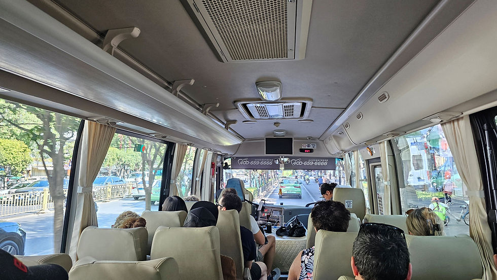 Bus interior with passengers seated, looking forward. Bright, sunny street visible through large windows. Overhead signs show phone numbers.