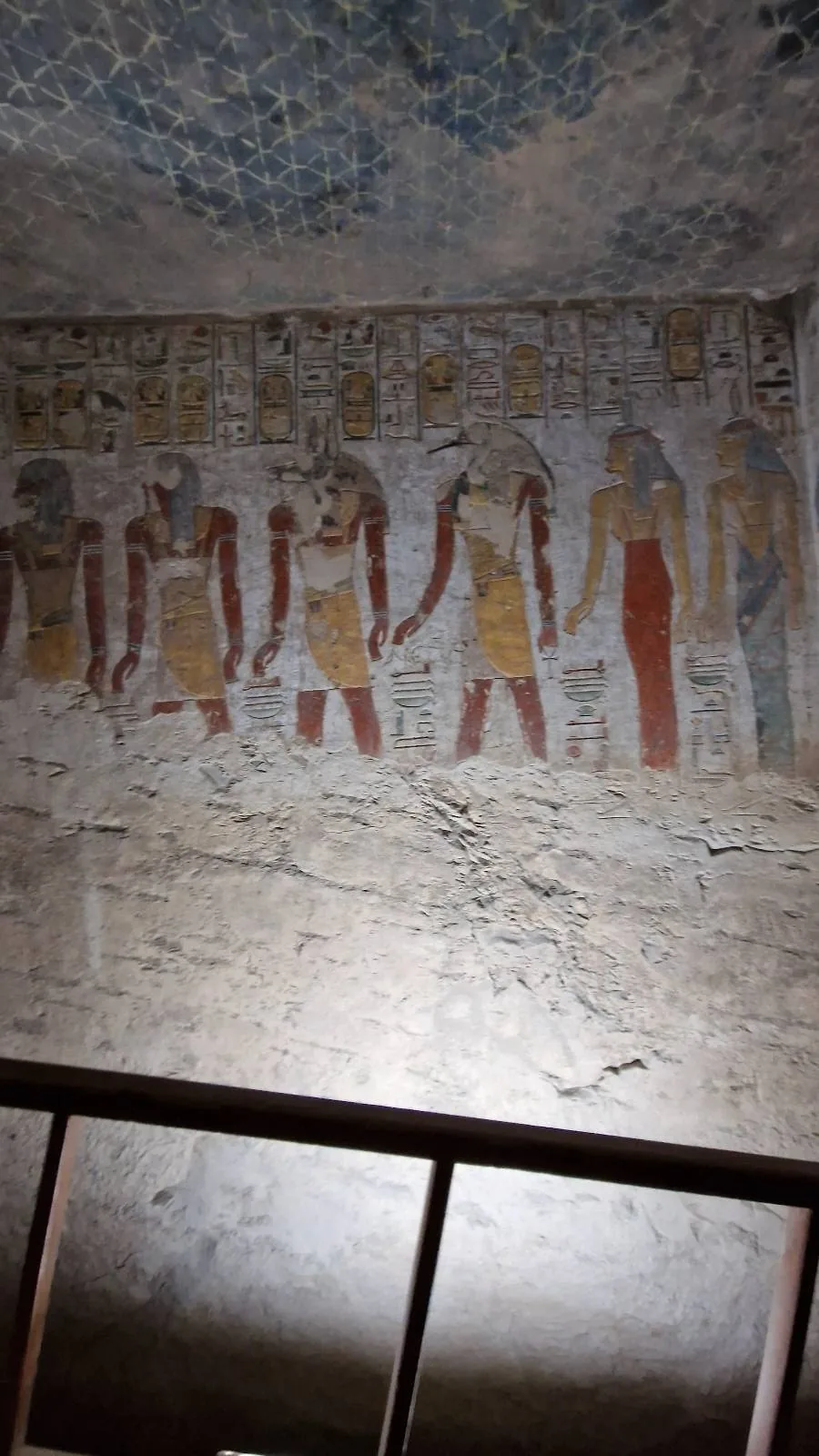 Ancient Egyptian wall painting depicting several figures in traditional attire, holding hands. The artwork includes hieroglyphics above the figures. The scene is illuminated by focused lighting, highlighting the details and colors of the mural.
