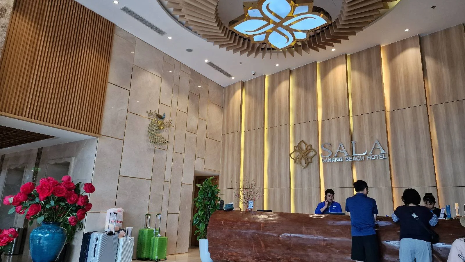 A hotel lobby with a wooden reception desk where two people are checking in. Behind the desk, two staff members in blue uniforms assist. The ceiling features a decorative light fixture, and the area is adorned with flowers and plants.