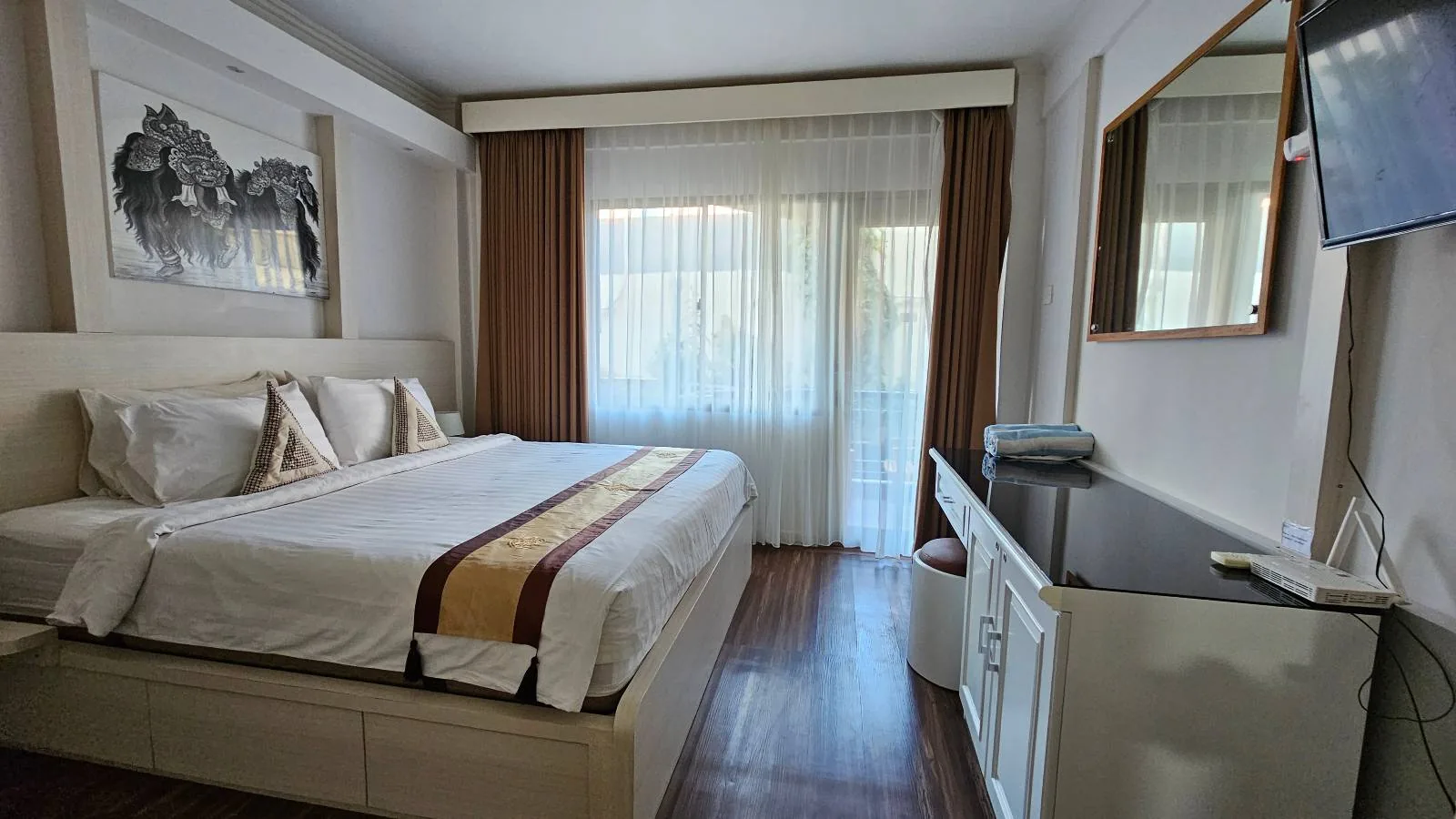 A modern hotel room with a large bed, white bedding, brown and yellow accent blanket, large window with sheer curtains, a wall-mounted TV, and a desk with a mirror.