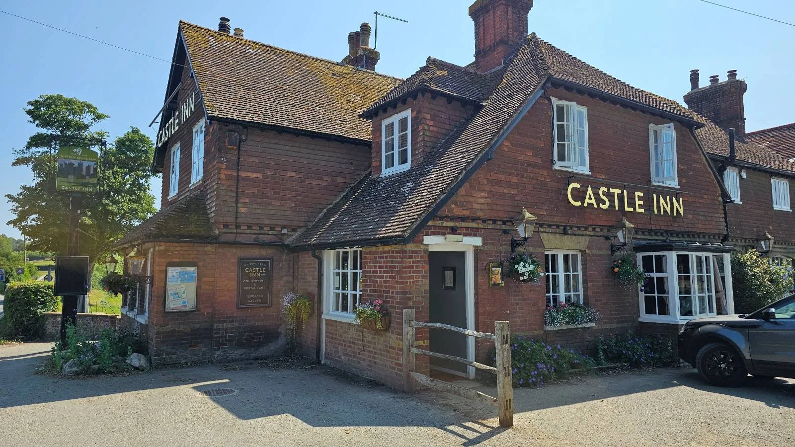 A traditional red-brick pub called "Castle Inn" with a sloped tiled roof, white-framed windows, hanging flower baskets, and a wooden sign, on a sunny day with blue sky.