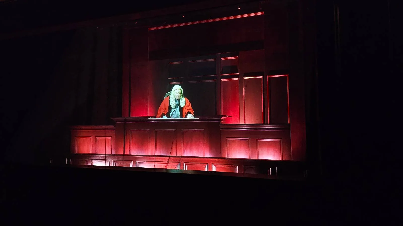 A person dressed as a judge sits behind a large, illuminated courtroom bench on a dark stage, lit by red and white lights.