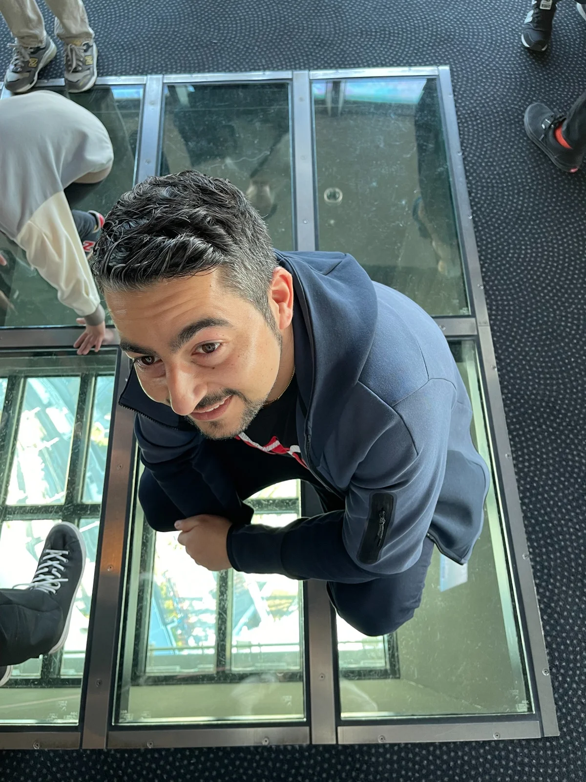 A person crouches on a clear glass floor, looking up at the camera with a slight smile. They are wearing a navy jacket and jeans. Other people's legs and shoes are visible around the person. The background shows a high view from the glass floor.