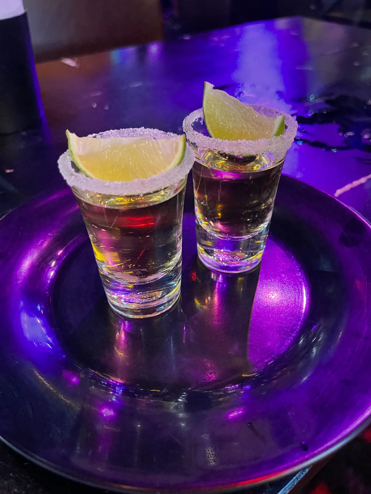Two shot glasses filled with clear liquid, each topped with a lime wedge, sit on a reflective dark plate under purple lighting.