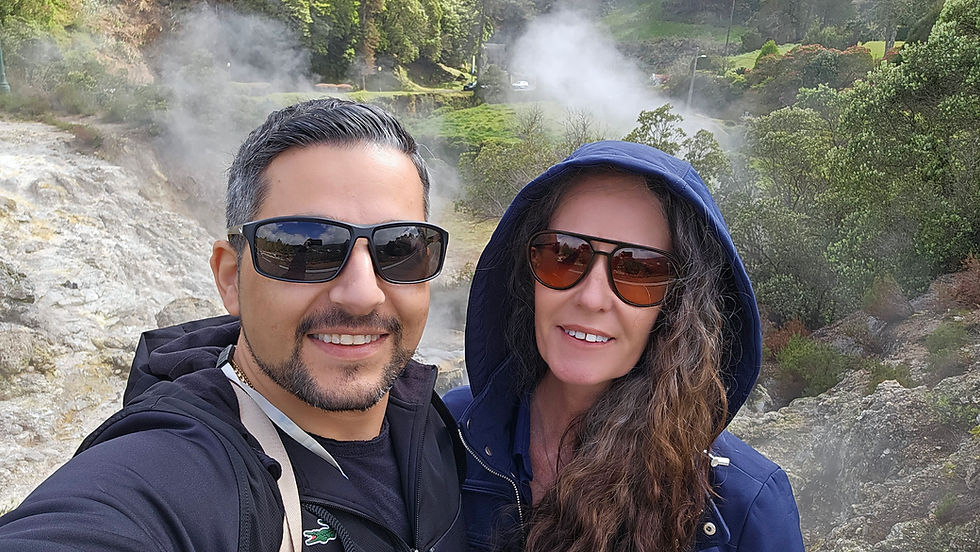 A man and woman wearing sunglasses and jackets smile for a selfie outdoors. Steam rises from the rocky, geothermal landscape behind them, with green trees and grass in the background.