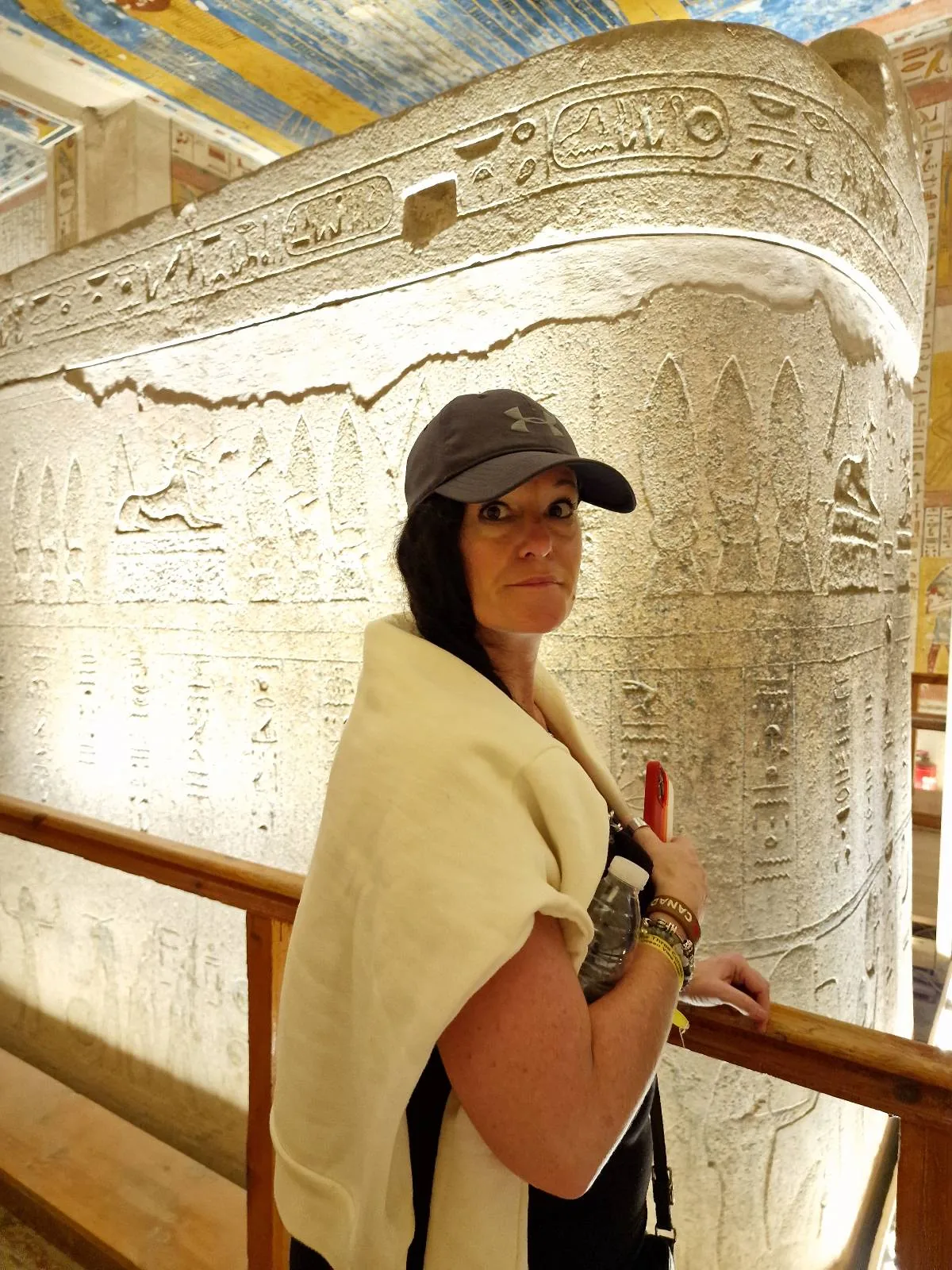 A person stands inside an ancient Egyptian structure, wearing a hat and holding a camera. They are in front of a large stone sarcophagus adorned with hieroglyphs. The ceiling features colorful painted designs.