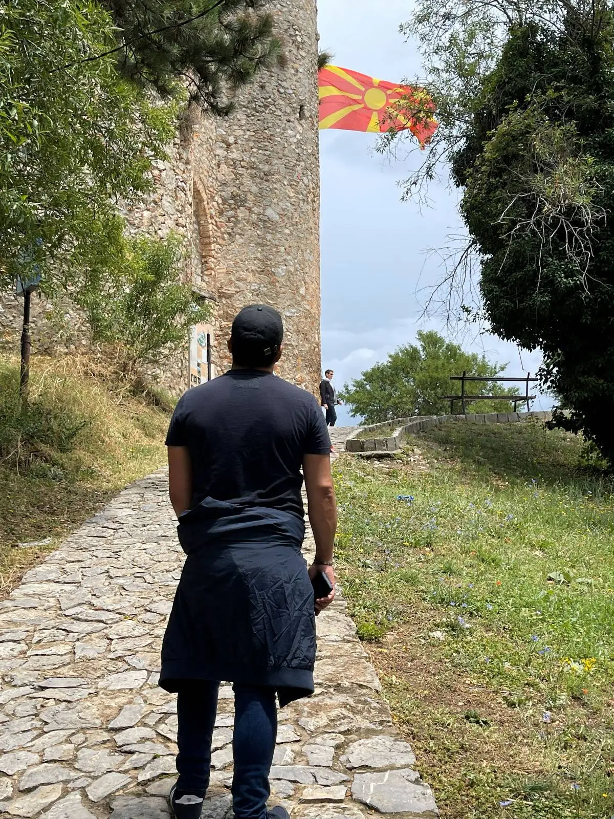 A person wearing a black shirt and cap walks along a stone path toward an ancient stone tower. A red and yellow flag with a sunburst design is flying near the top. The scene is surrounded by green trees and grass under a cloudy sky.