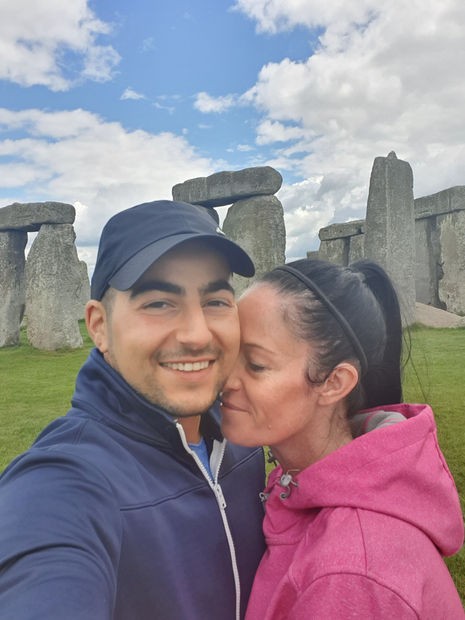 A couple poses smiling for a selfie in front of the ancient monument of Stonehenge. The man sports a navy cap and jacket, while the woman in a pink hoodie leans her head affectionately against his cheek. The sky above the English countryside is partly cloudy.