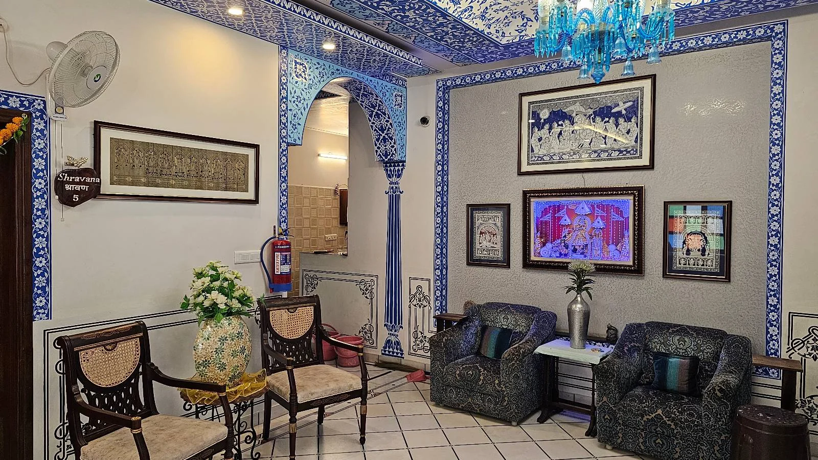 A cozy living room with ornate blue and white decor, patterned ceiling, framed art on the walls, two dark sofas, two wooden chairs, a blue chandelier, and a small table with flowers.