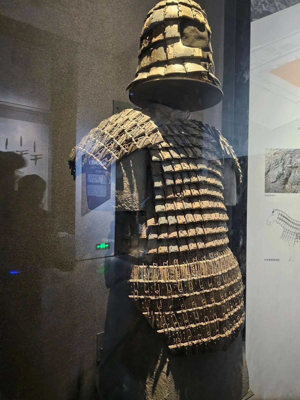 A display of ancient segmented armor with overlapping metal plates, including a helmet and protective gear for the shoulders and torso, exhibited in a museum setting behind glass.